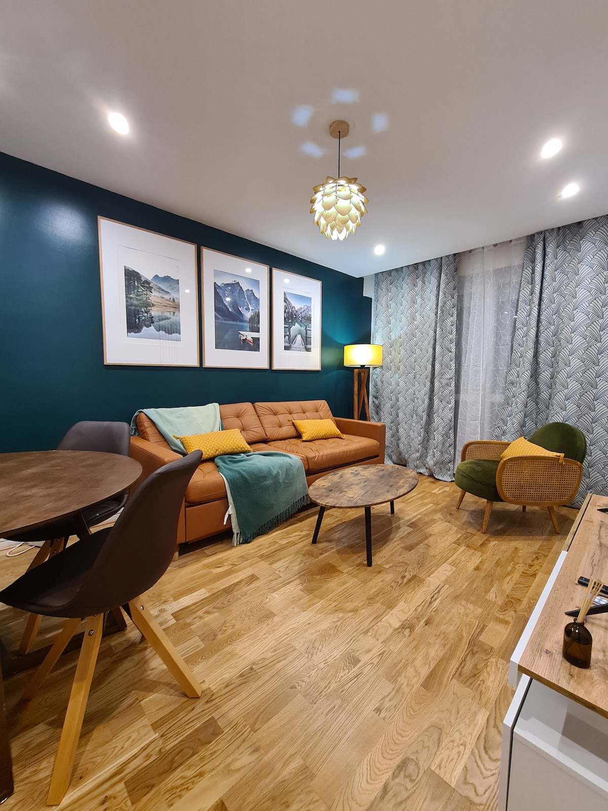 A tastefully decorated living area features a brown sofa adorned with cushions, complemented by a small round table and a green accent chair. Large windows are draped with patterned curtains, and framed photographs hang on the walls, enhancing the warm ambiance.
