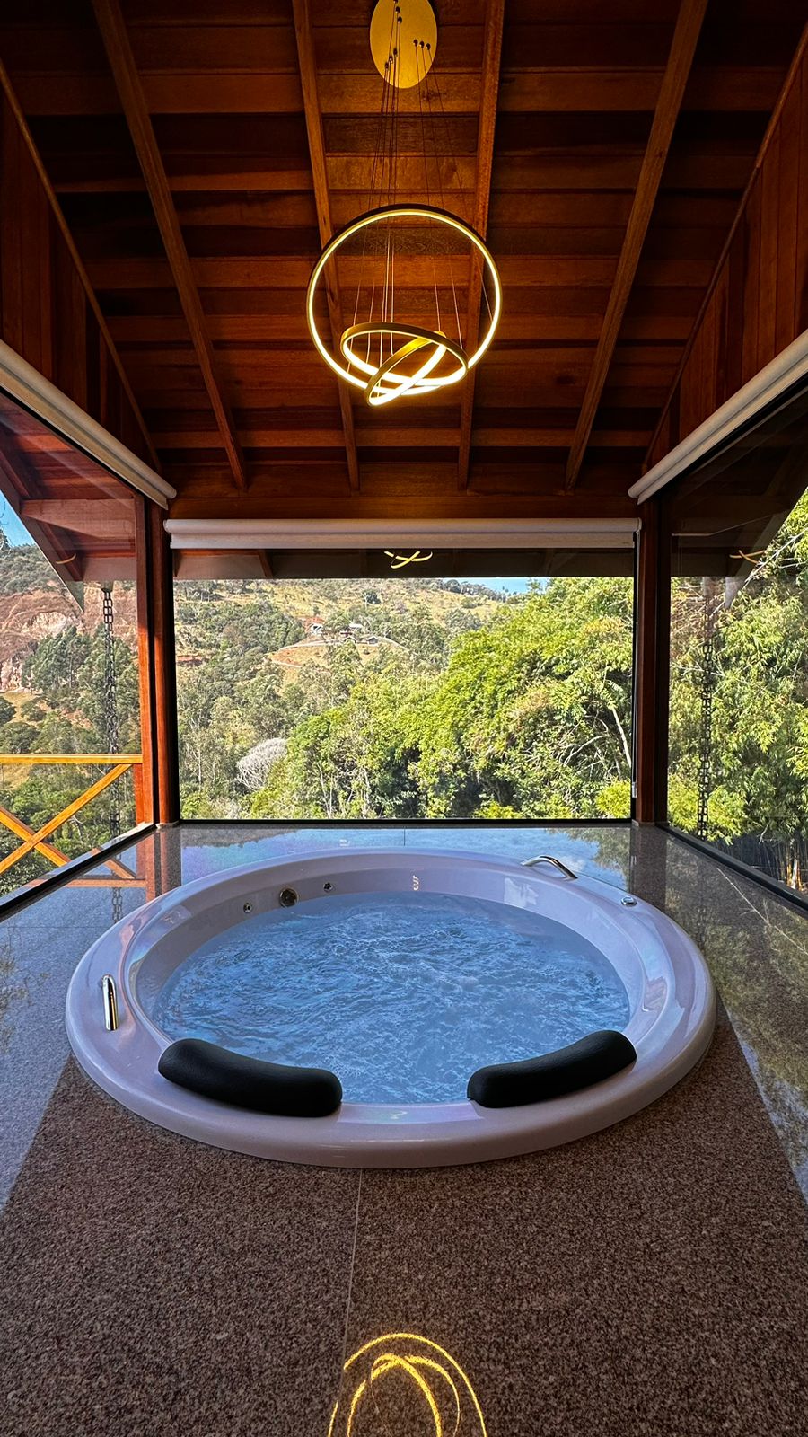 A private hot tub is situated on a polished stone surface, offering panoramic views of the lush green landscape through floor-to-ceiling glass windows. A modern light fixture hangs above, complementing the rustic wooden ceiling.