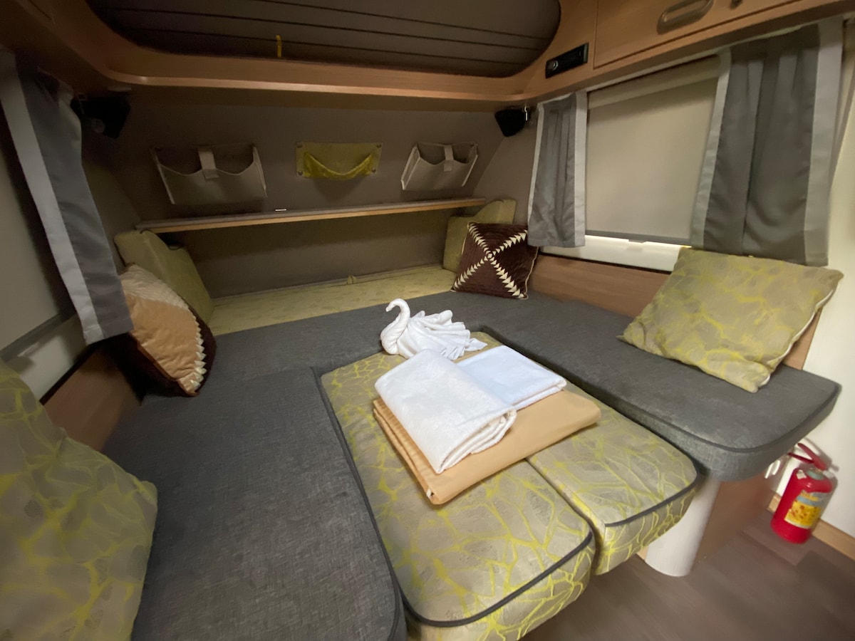A compact interior of the caravan is presented with a spacious seating area featuring soft grey and patterned cushions. A neatly arranged wooden table holds folded towels and a book. Above are storage pockets, with natural light filtering through the windows, dressed in grey curtains.