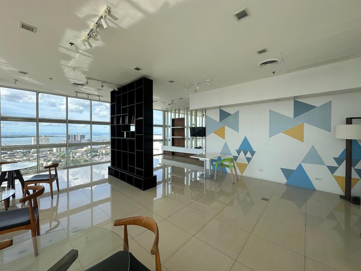 An open living space features large windows offering views of the ocean and cityscape. Light-colored tiles reflect natural light. A modern seating area with wooden chairs is complemented by a stylish black feature wall, while geometric patterns adorn the adjacent wall.