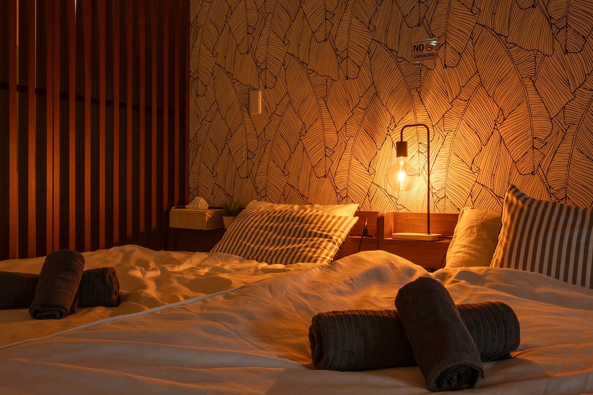 A softly lit bedroom features two spacious beds adorned with neatly rolled towels. The walls are decorated with a nature-inspired pattern, and a warm-toned bedside lamp casts a gentle glow, enhancing the tranquil ambiance.