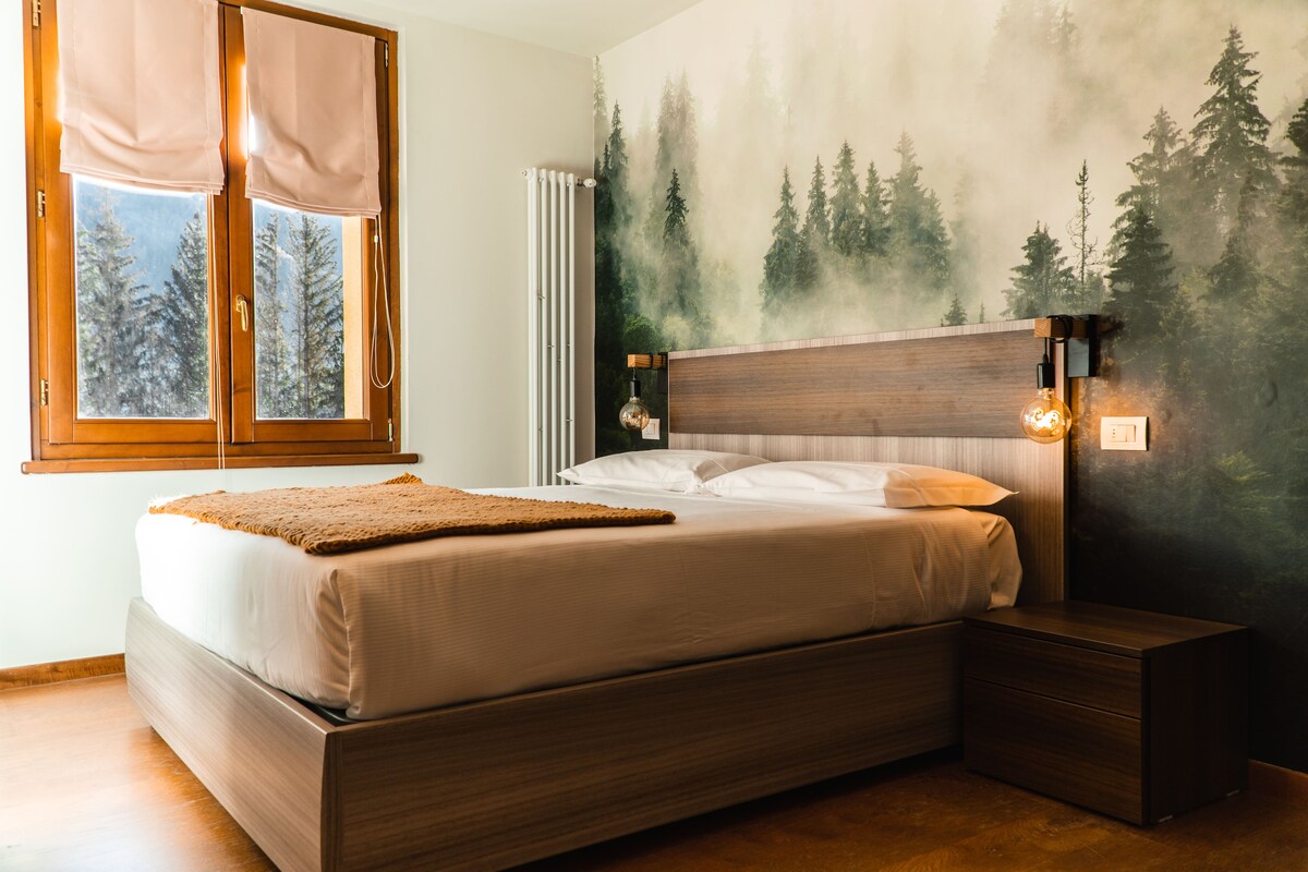 A serene bedroom is adorned with a mural of dense forest trees, creating a tranquil atmosphere. The spacious bed is accompanied by a bedside table with a soft-textured throw draped over it. Two large windows allow natural light to filter in softly.