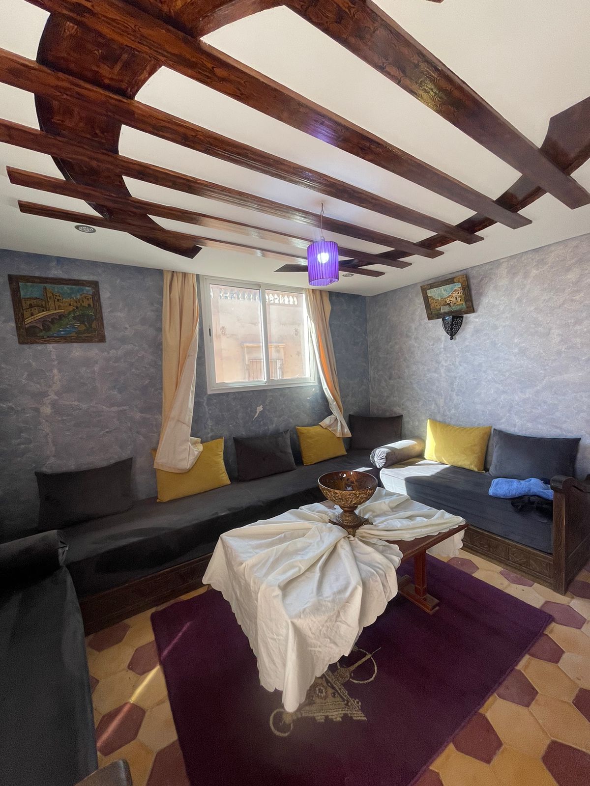 A cozy seating area features low, dark couches adorned with yellow accent pillows. A wooden table sits in the center, topped with a decorative bowl. Natural light enters through a window, complemented by a ceiling fan and textured walls with artistic decorations.