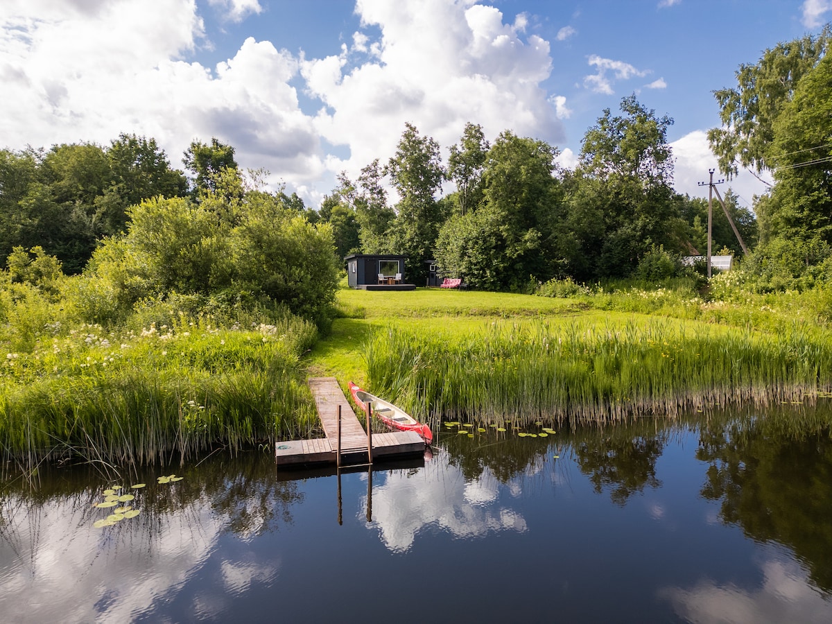 A peaceful natural setting is depicted, featuring a wooden dock extending into a calm body of water. Lush greenery surrounds the area, with tall grasses and trees visible. The minimaja can be seen in the background, nestled among the trees under a partly cloudy sky.