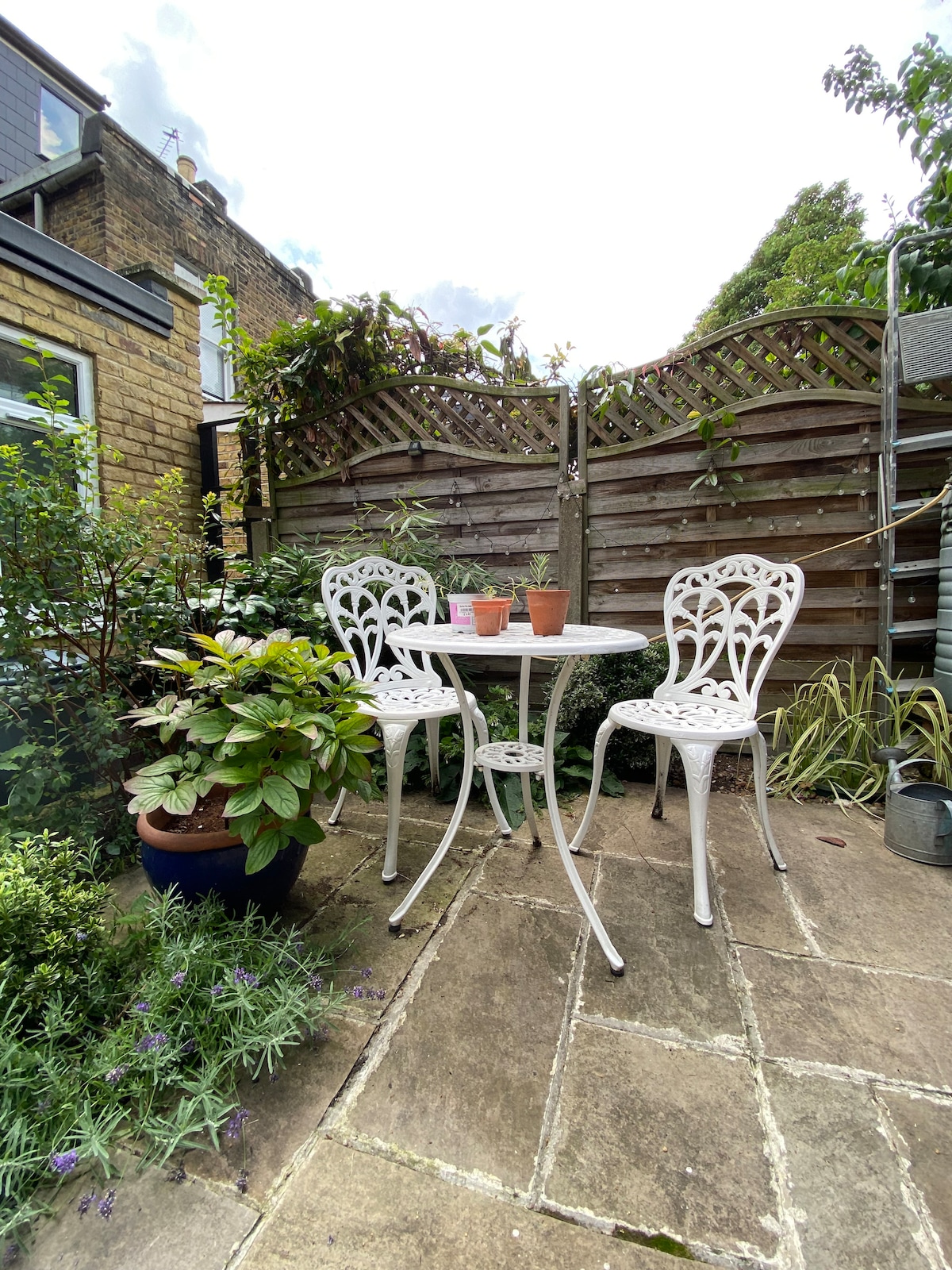 A quaint garden area features a white metal table surrounded by two chairs. Potted plants sit on the table, with lush greenery and flowering plants creating a serene backdrop. The stone patio is laid out in a rectangular pattern, enhancing the outdoor setting.