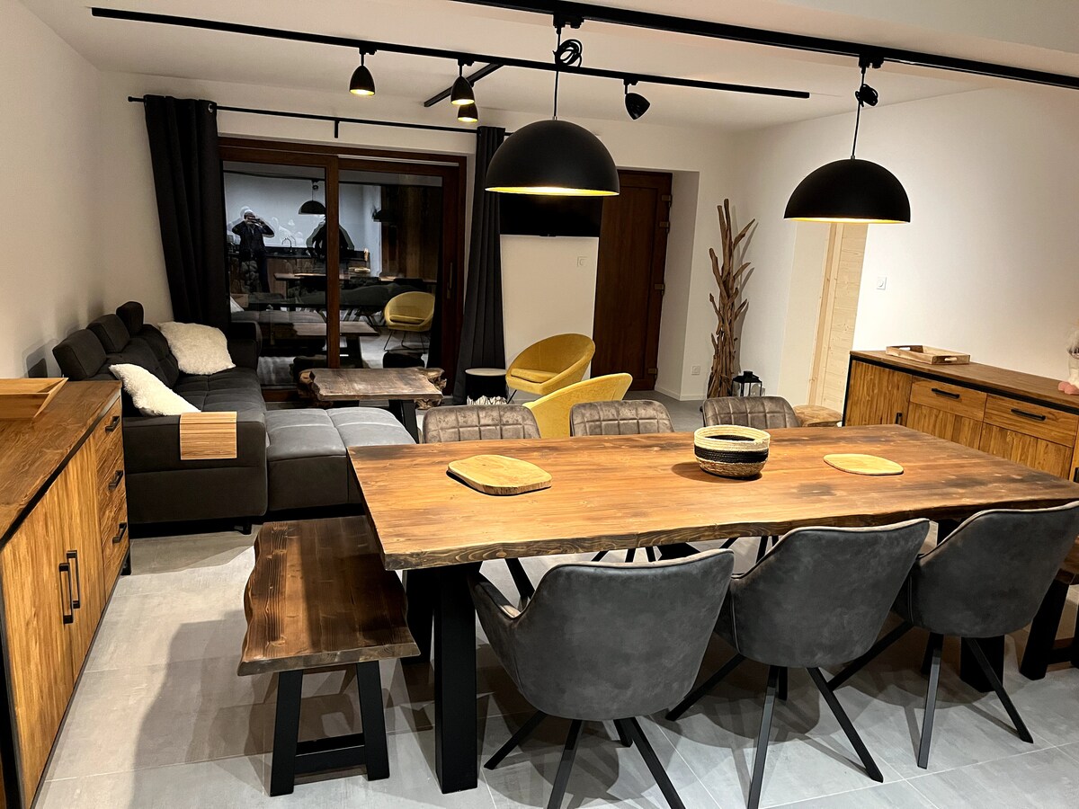 A spacious dining area is highlighted with a large wooden table surrounded by six upholstered chairs. A comfortable sofa and yellow accent chair are visible in the background. Simple yet elegant lighting fixtures hang above, and large windows allow for natural light to fill the room.