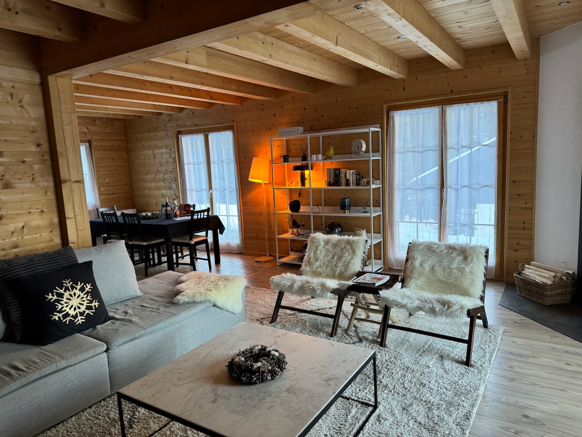 The living area features a spacious sectional sofa and two lounge chairs, all situated on a plush area rug. A rustic dining table is visible, accompanied by black wooden chairs. Natural light enters through large windows, and a warm lamp casts a gentle glow.