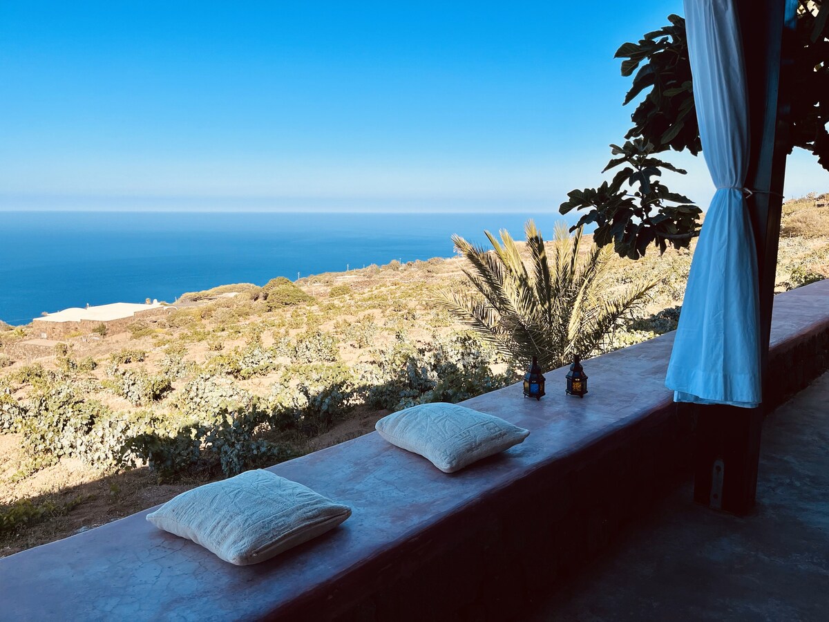 A serene outdoor terrace features three soft cushions arranged on a ledge, with two small lanterns for ambient lighting. The view stretches across lush landscapes towards the horizon, meeting a clear blue sky and the sea beyond.
