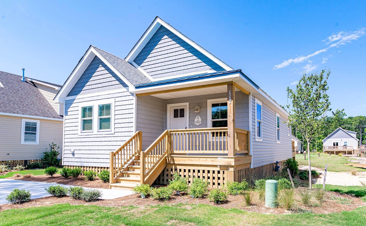 The cottage presents a welcoming facade with light grey siding and a covered front porch. A wooden staircase leads to the entrance, surrounded by well-maintained landscaping featuring low shrubs and fresh green grass. The exterior reflects a charming and modern architectural style.