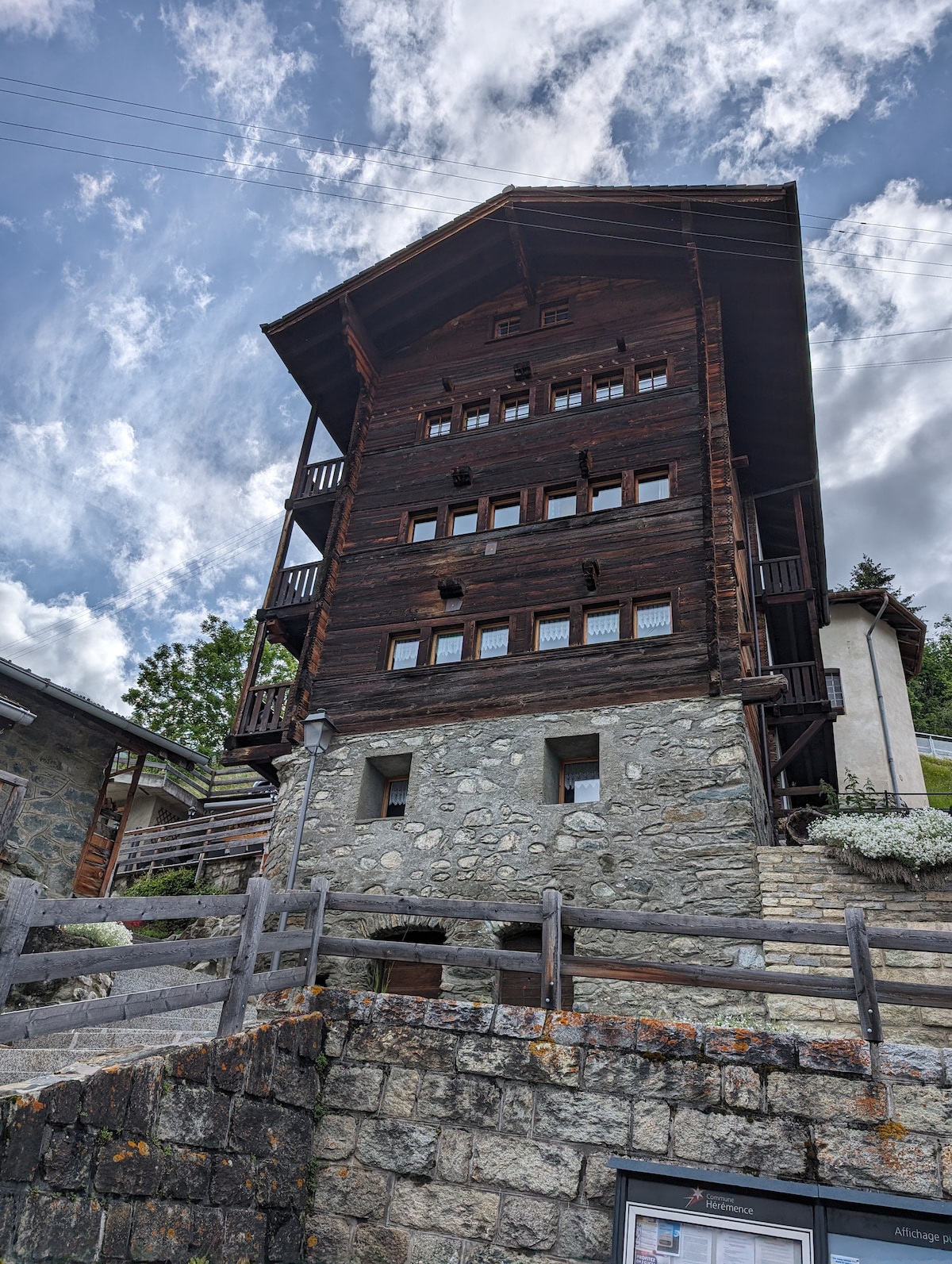 A historic chalet-style building with a combination of wood and stone construction is shown in the image. The structure features multiple balconies, large windows, and a sloped roof. The surrounding area includes a wooden fence and greenery, reflecting its setting in a mountainous village.