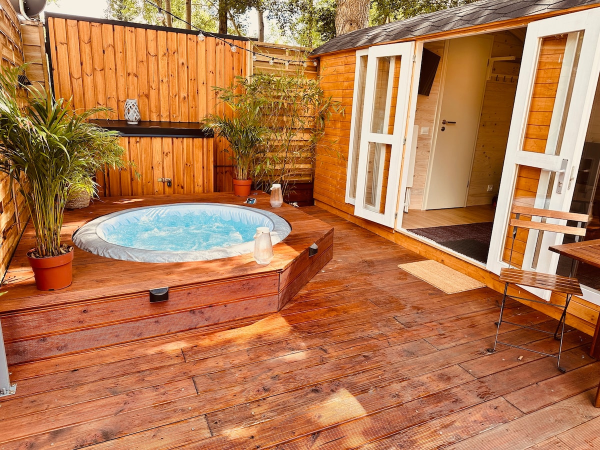 An outdoor wooden deck features a private whirlpool surrounded by potted plants. The entrance to the Tiny House is visible, with double glass doors providing access. The warm wood tones of the deck create a natural and calming space for relaxation.