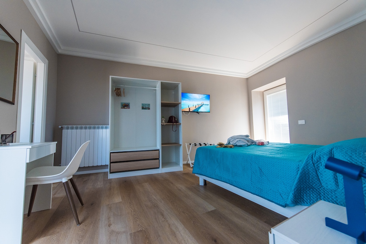 A spacious bedroom is featured with a vibrant blue bedspread covering the bed. A wall-mounted TV is visible, alongside a wardrobe and a desk with a chair. Light streams in through the window, highlighting the warm wooden flooring.