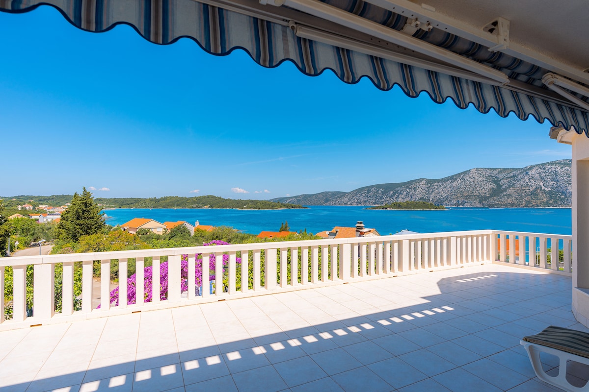 A spacious terrace offers a panoramic view of the clear blue Adriatic Sea and the lush hills of the Pelješac Peninsula. Bright sunshine floods the area, enhancing the light-colored tiles and the crisp white railing, while a striped awning provides unobtrusive shade.