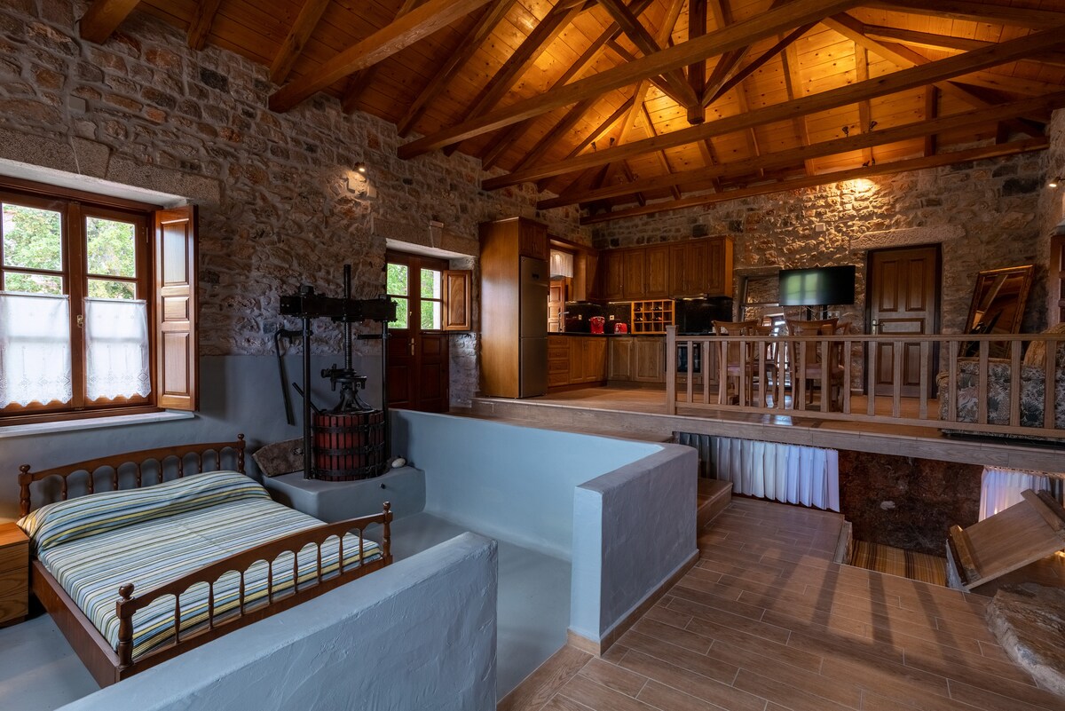 The interior features a blend of stone walls and wooden beams, showcasing an open-plan space. A cozy sleeping area with a striped bedspread is situated on a raised platform, while a traditional winemaking press adds historical charm. The kitchen area is visible, well-equipped for guest needs.