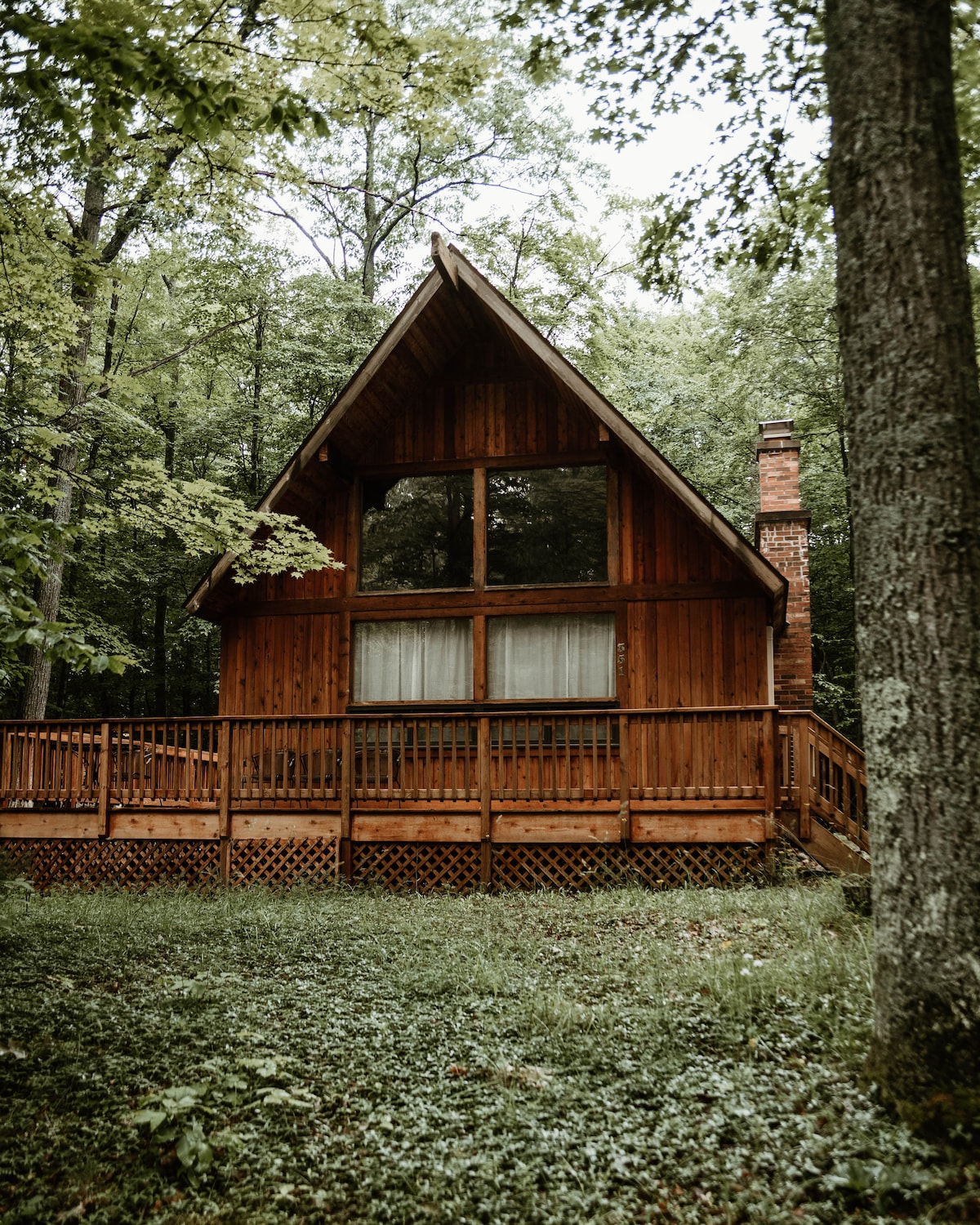 The chalet features a distinctive A-frame architecture, with wooden siding and a spacious deck surrounded by lush greenery. Large windows allow for natural light to fill the interior, while a stone chimney adds character to the rustic exterior.