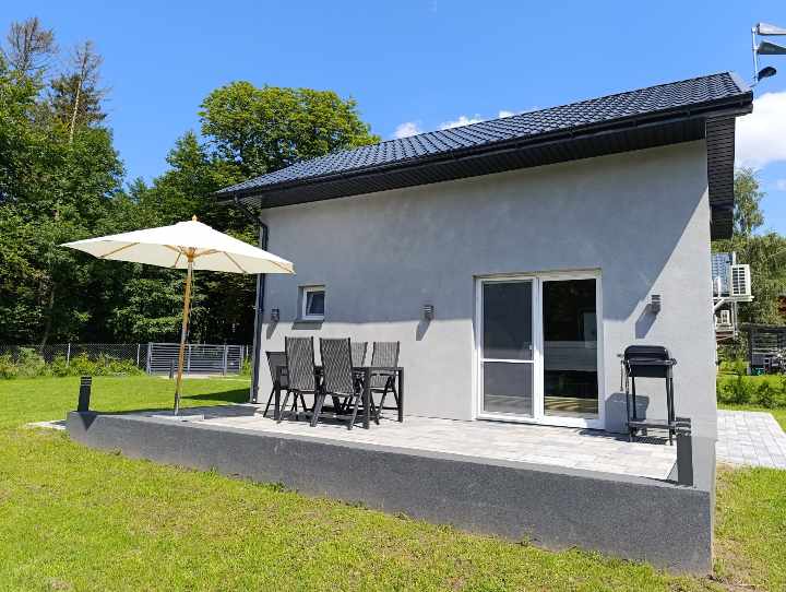 A modern building features a spacious outdoor terrace with a large umbrella providing shade. A dining set for six is visible, accompanied by a grill. The well-maintained surrounding garden and trees create a peaceful retreat setting.