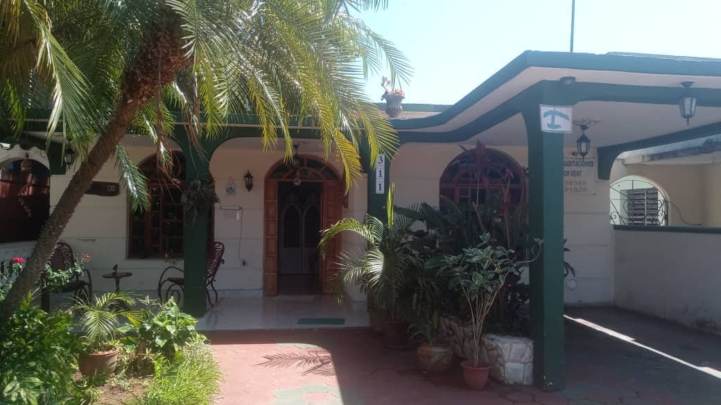 A welcoming entrance features lush tropical plants and palm trees, with a shaded patio showcasing decorative wrought iron furniture. The facade has arched windows and a wooden door framed by greenery. The pathway is paved, leading to the inviting interior of the casita.