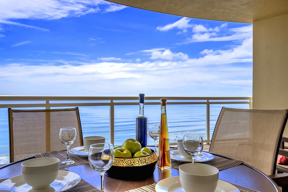 A dining table is set for six, featuring white bowls and crystal glasses. A decorative bowl of green apples sits at the center, complemented by bottles of oil and vinegar. The balcony offers a panoramic ocean view under a clear blue sky.