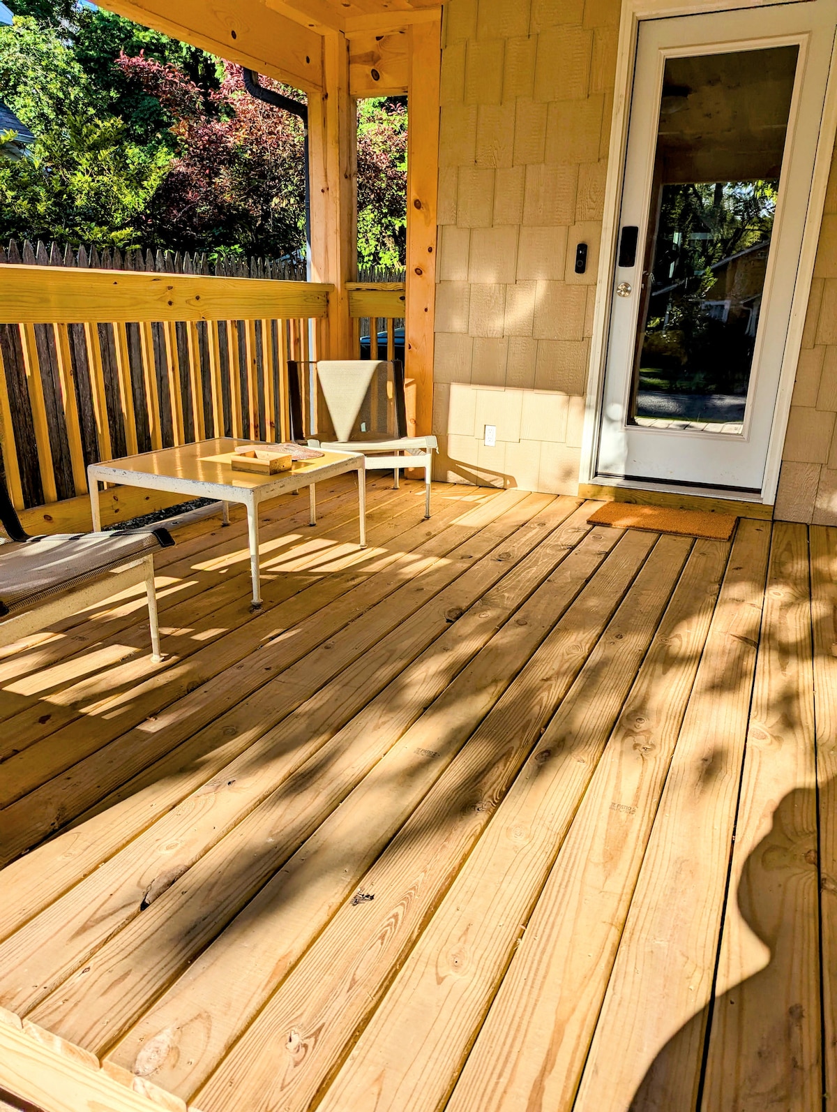 A covered porch area features a wooden floor, with sunlight casting shadows. Two light-colored chairs are positioned around a small table. A glass door leads to the interior, framed by wooden siding and surrounded by greenery.