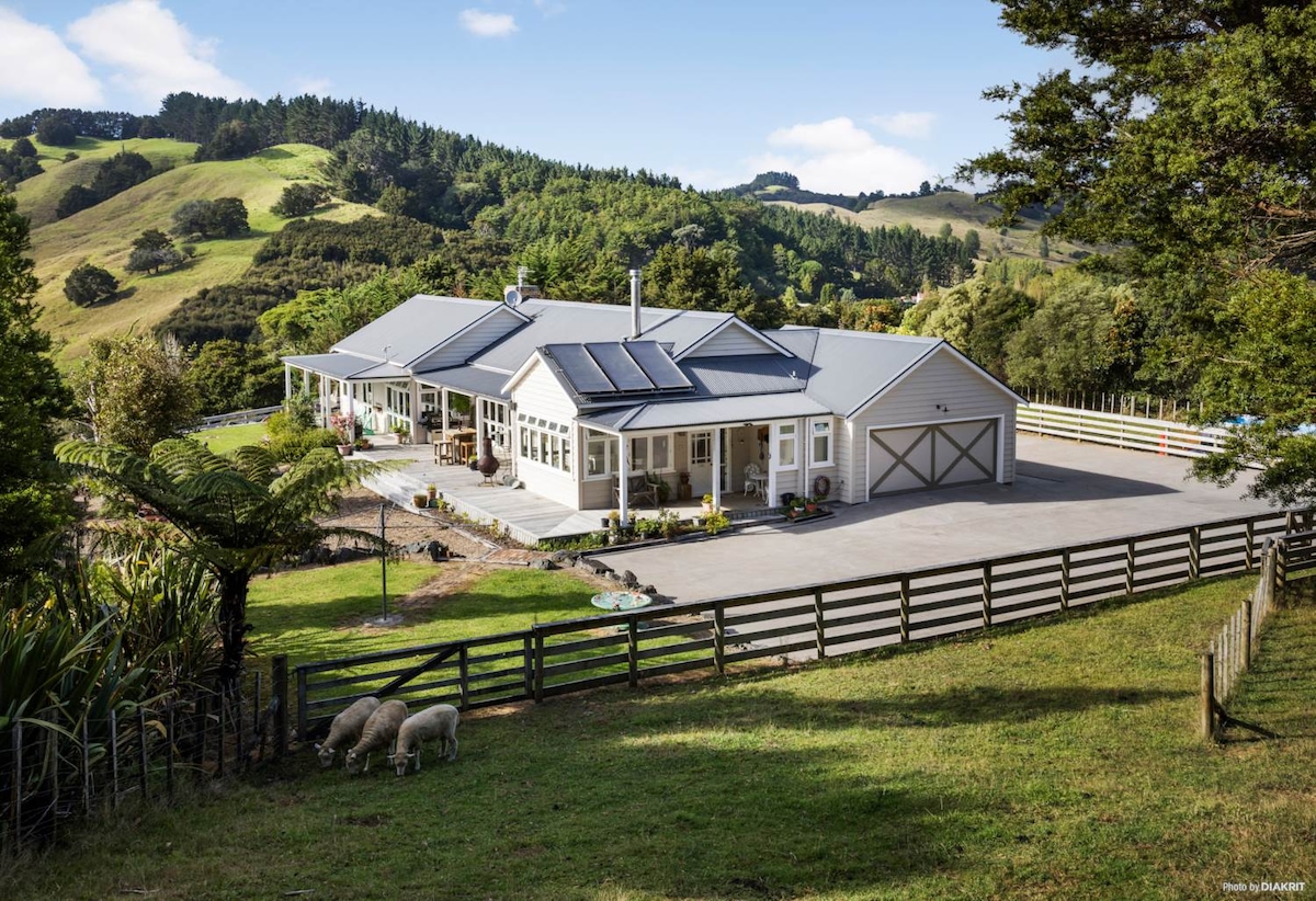 The spacious farmhouse is set against a backdrop of lush green hills. Large windows reflect natural light, while a wide porch invites relaxation. A grazing area is visible in the foreground, showcasing sheep amidst the landscaped surroundings, highlighting the property’s harmonious connection with nature.