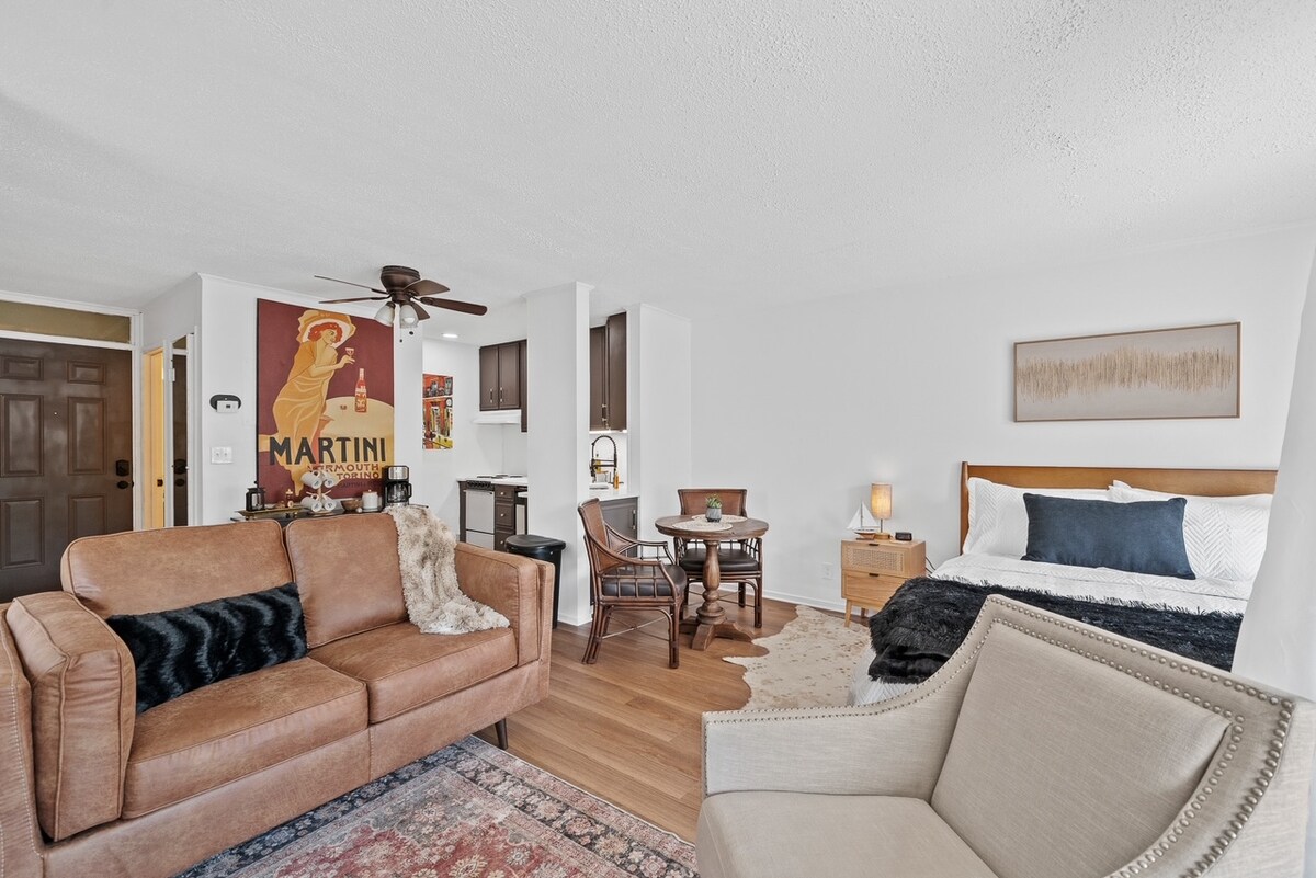 A studio space features a cozy seating area with a brown leather sofa and an armchair. The bed is positioned near a wall adorned with a vintage martini poster. A small dining table is seen beside the bed, while the kitchen area is visible in the background.