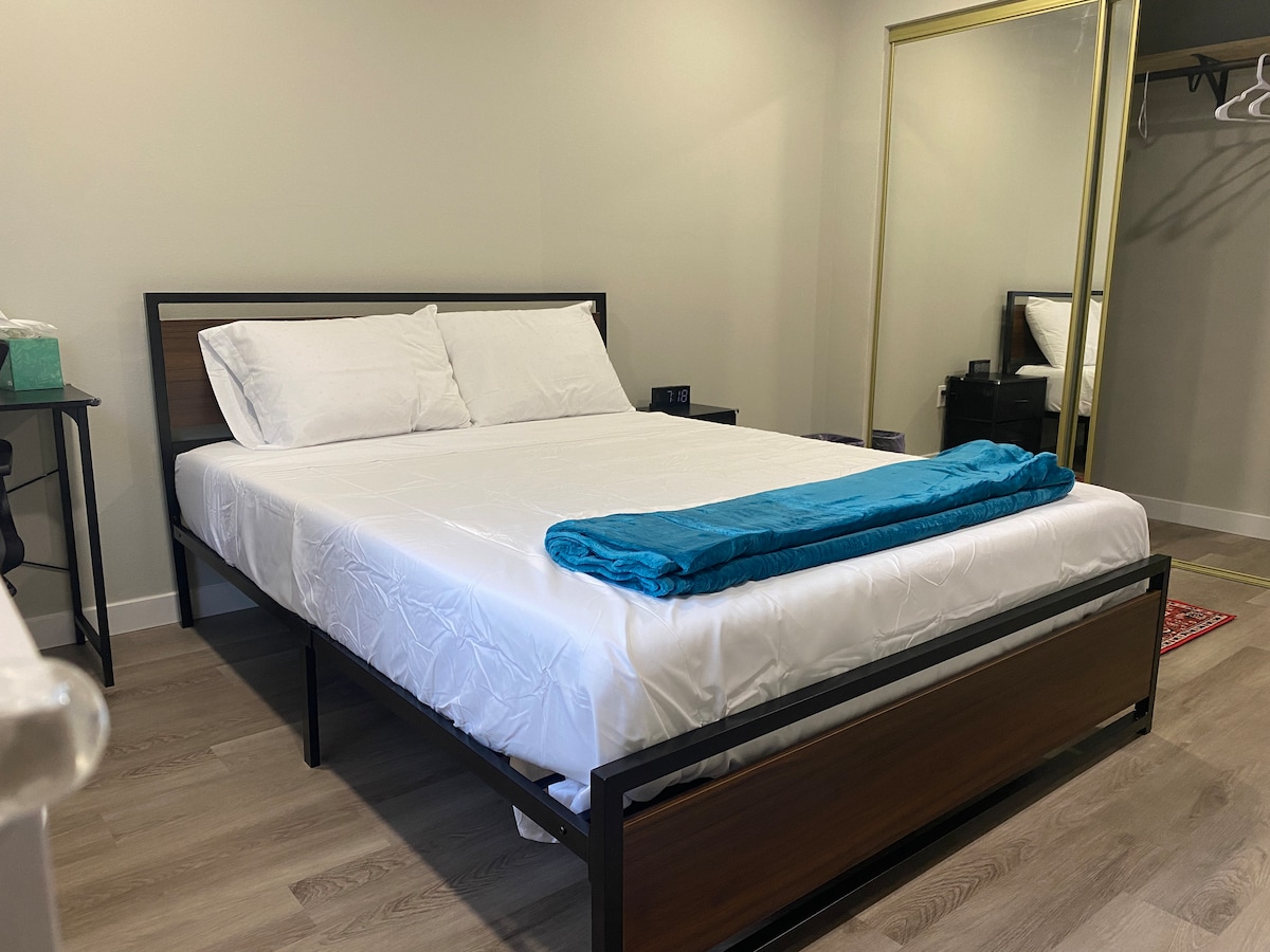 A spacious room features a queen bed with white bedding and a teal throw blanket, positioned against a minimalist backdrop. A mirrored closet door reflects the room, while a small table with a clock is placed next to the bed. Wooden flooring adds warmth to the space.
