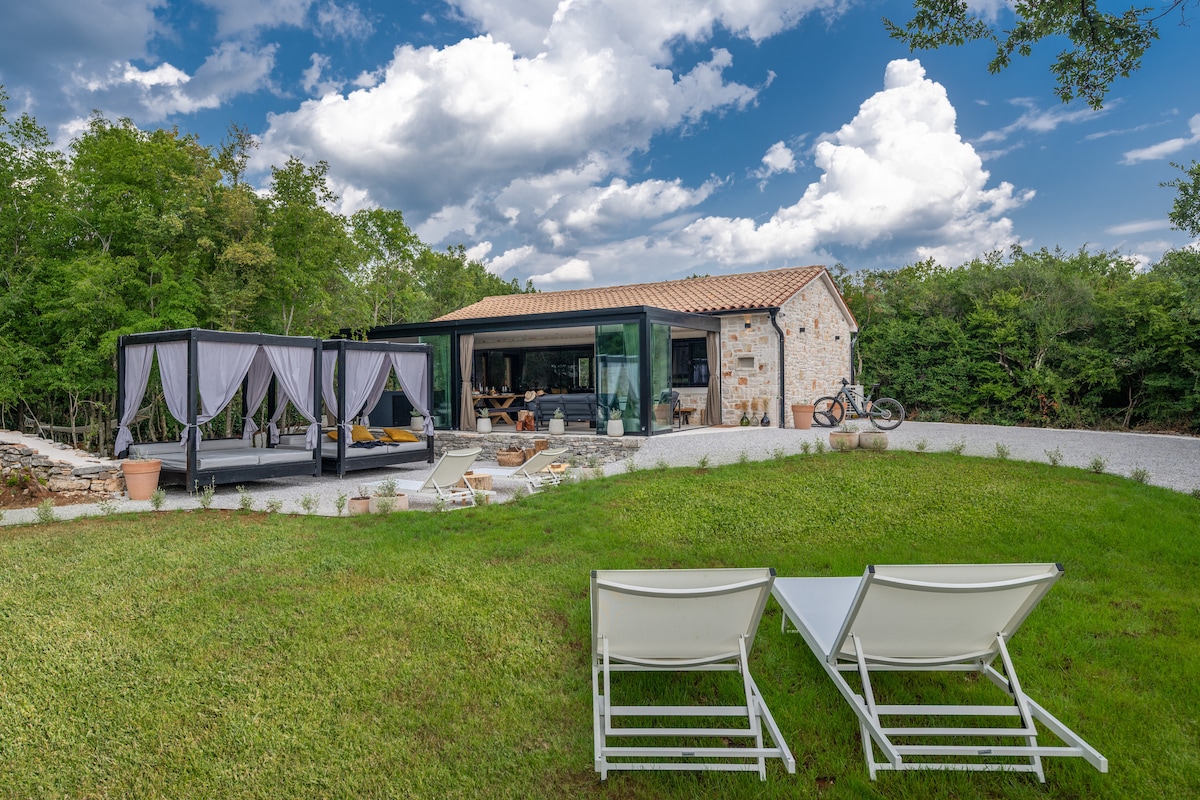 A charming house constructed with traditional stone elements is surrounded by lush greenery. A large terrace is visible, featuring elegant drapes and a comfortable seating area. Two lounge chairs are positioned on the manicured lawn, offering a relaxing space to enjoy the tranquil outdoor setting.