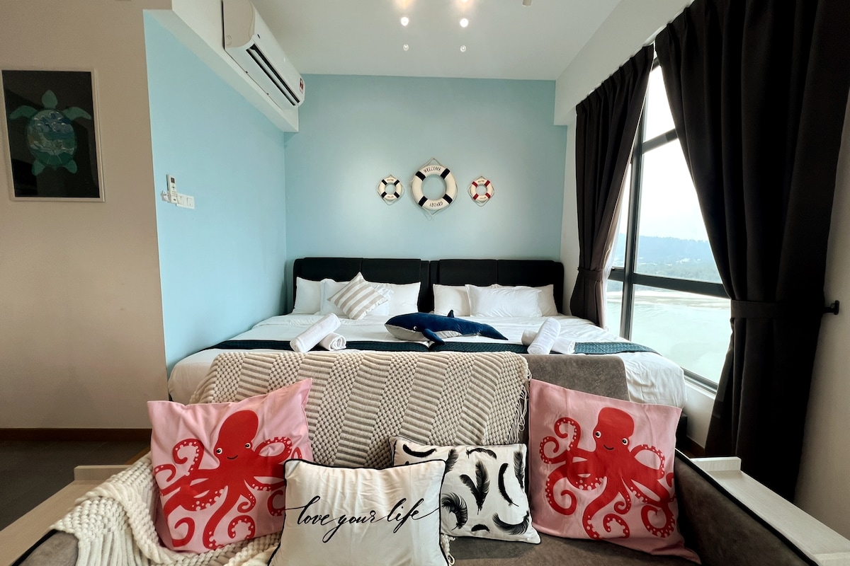 A well-appointed room features two spacious beds with soft linens, set against a soothing blue wall. Decorative pillows with octopus designs adorn the seating area, while large windows offer ample natural light and a view of the surroundings.
