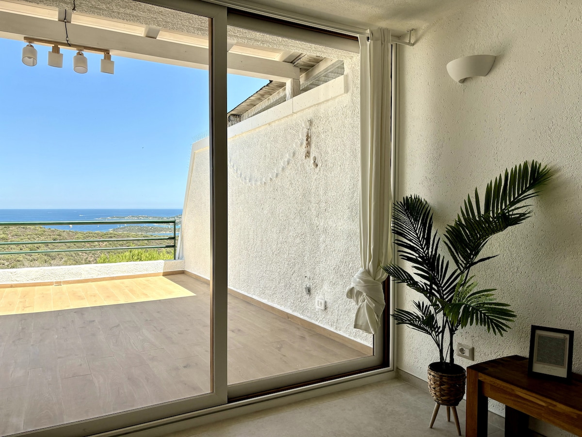 A spacious living area contains large glass doors that open to a terrace with a sea view. Natural light floods the room, highlighting a green potted plant and a wooden side table. The neutral-toned walls and flooring contribute to a calm environment.