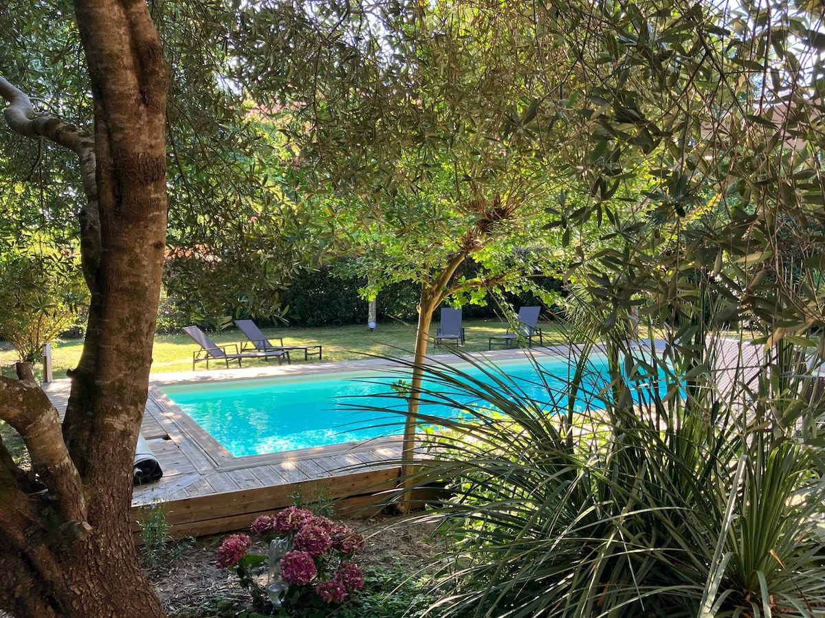 A private swimming pool is surrounded by a wooden deck, with several lounge chairs positioned nearby. Lush greenery frames the area, providing shade and privacy. Colorful flowers are visible in the foreground, adding to the serene garden setting.