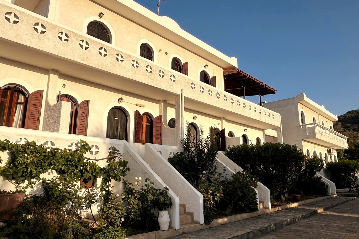 A stunning whitewashed building features multiple arched windows and wooden shutters. The structure is accented by decorative details and is surrounded by lush greenery. Stone steps lead up to the entrance, enhancing the inviting atmosphere of the property.