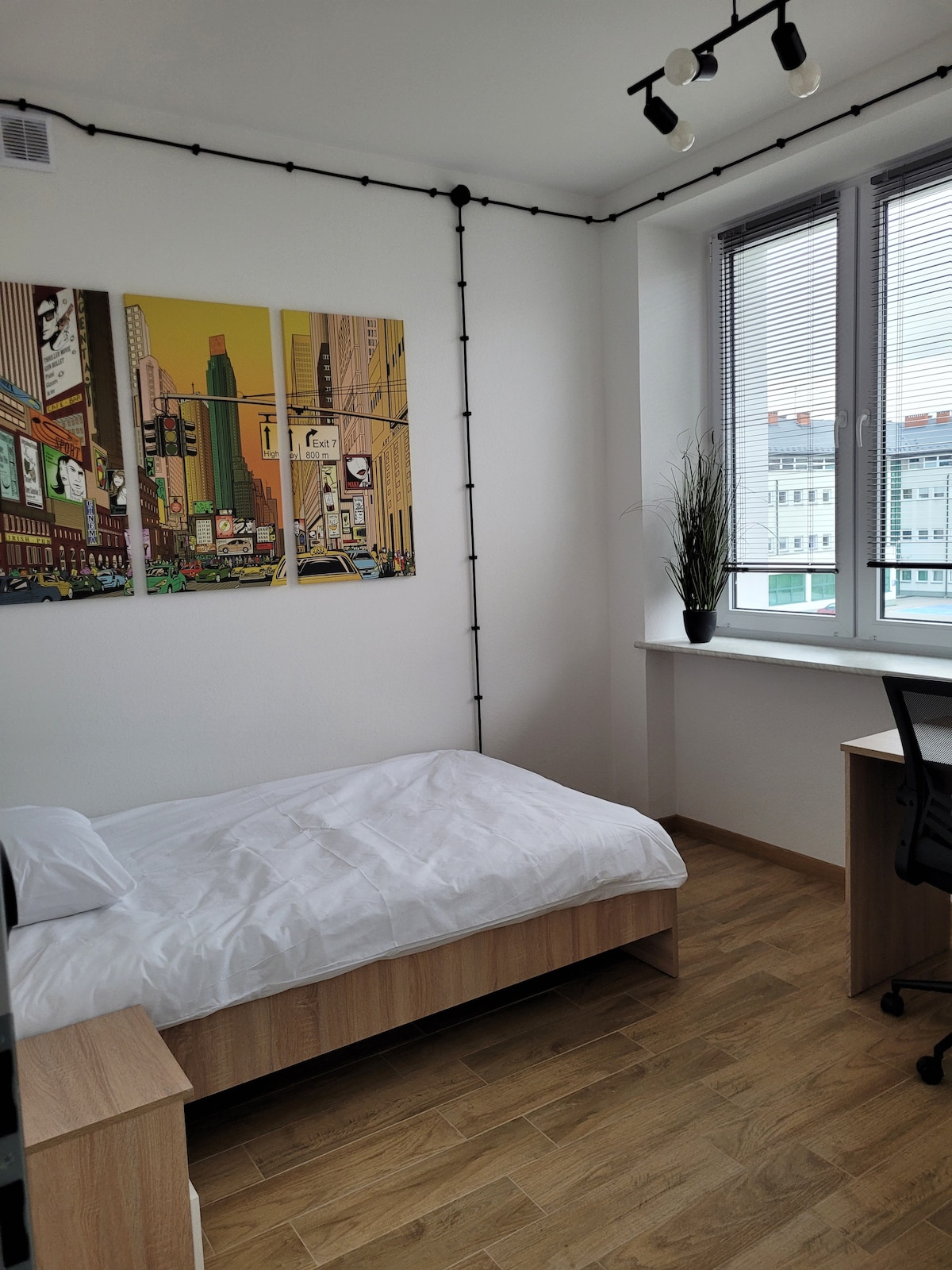A bedroom features a spacious bed with white linens, complemented by light wood furniture. A desk and chair are positioned near a window that allows natural light to fill the space. Colorful artwork depicting a cityscape adorns the wall.