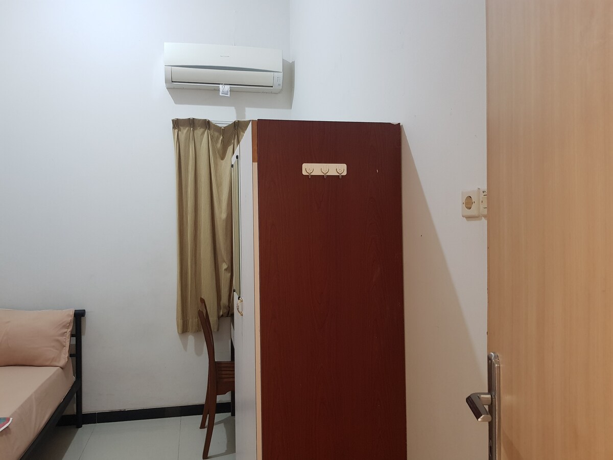 A simple room is depicted featuring a single bed and a wooden wardrobe. A chair is positioned nearby, and a small window covered by light curtains allows natural light to enter. An air conditioning unit is mounted on the wall.