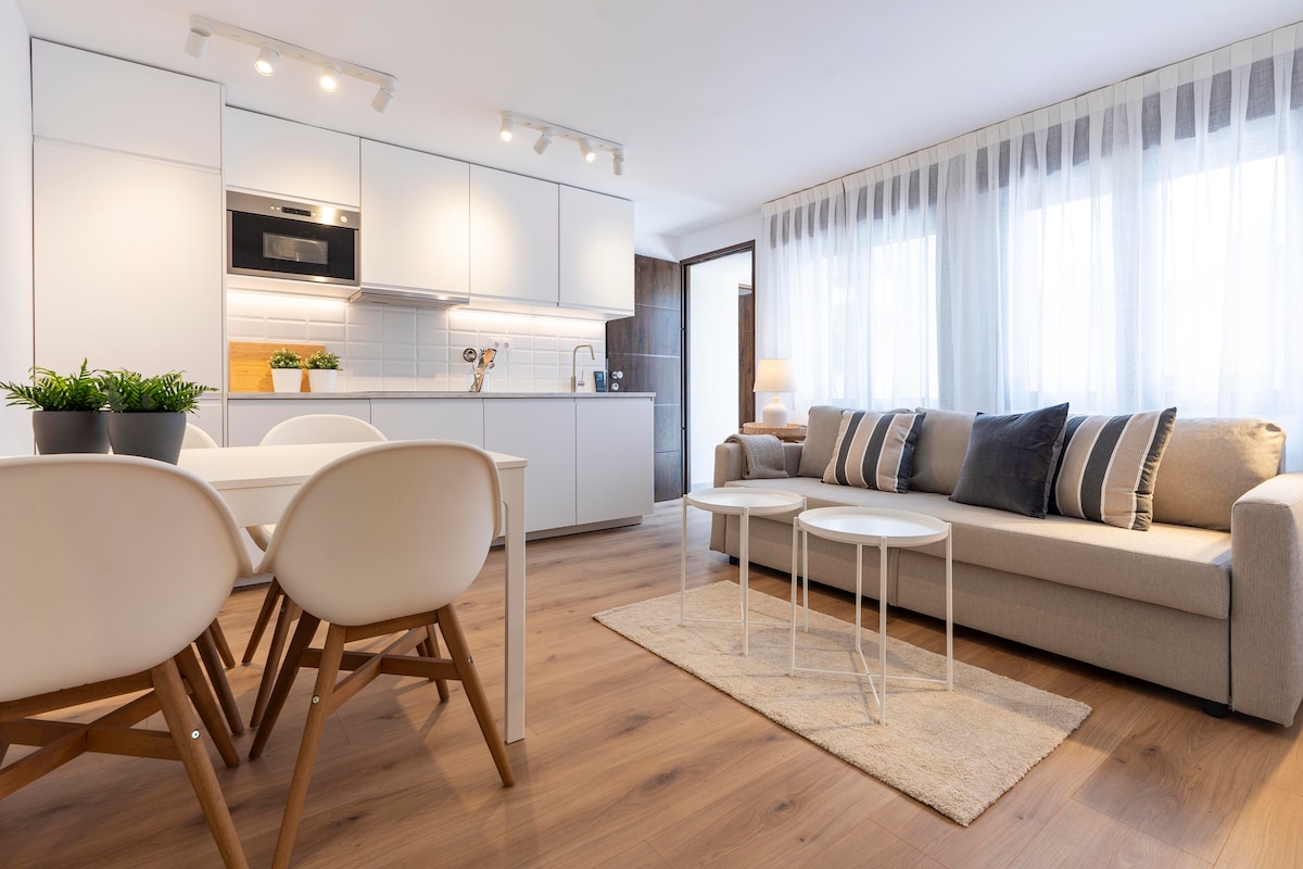 A modern living space features a cozy sofa complemented by decorative cushions. A dining table with four chairs is positioned beside a well-equipped kitchen with sleek cabinetry. Natural light enters through large windows, enhancing the inviting atmosphere of the room.