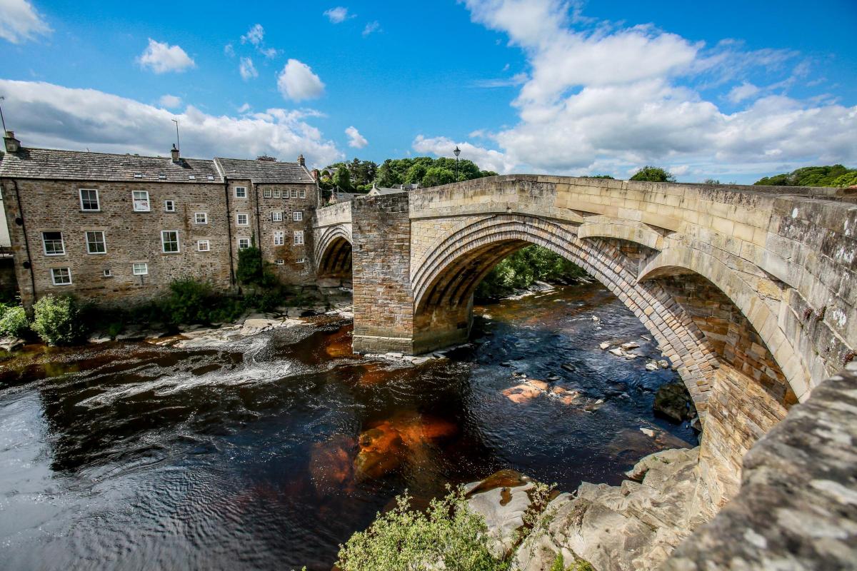 A historic stone bridge spans a river, with its arches gracefully reflected in the water. Lush greenery lines the riverbank, and traditional buildings are visible in the background under a partly cloudy sky.