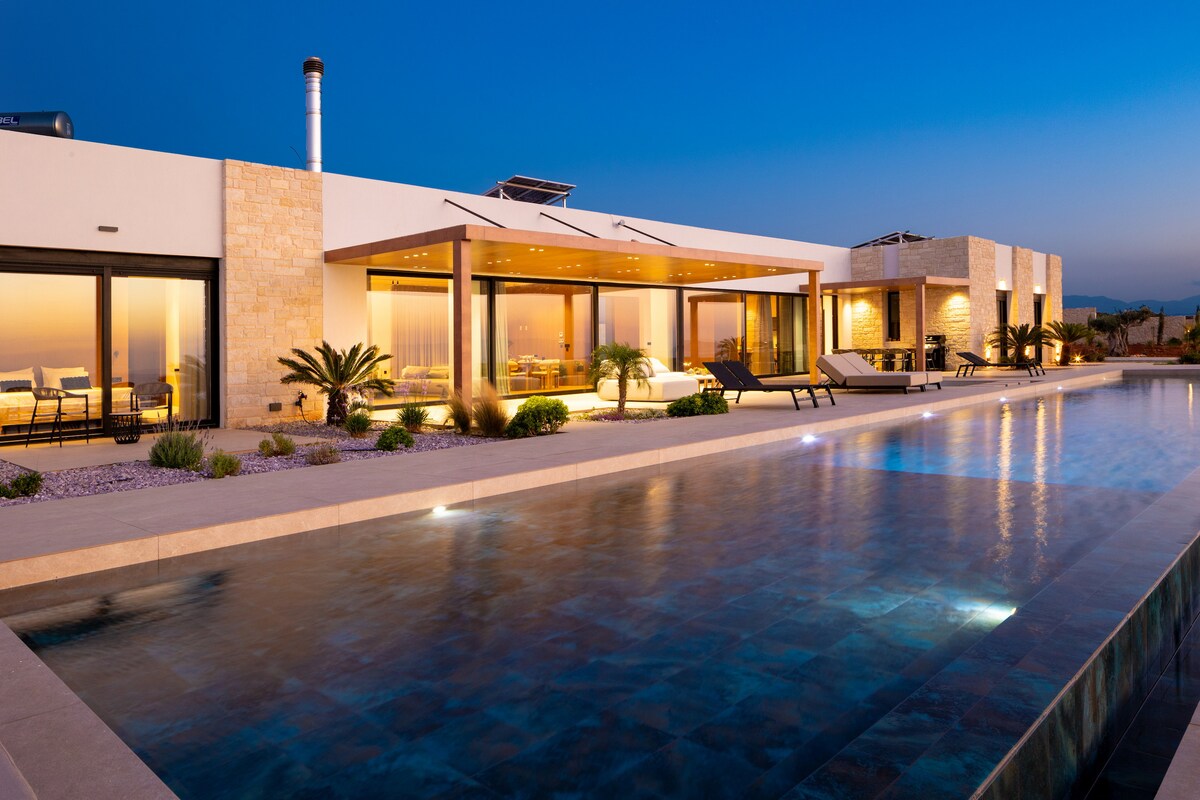 An elegant single-story villa is illuminated at dusk, featuring a spacious swimming pool reflecting soft lights. Floor-to-ceiling windows provide views of the inviting interior spaces, highlighting a blend of comfort and contemporary design. Surrounding landscaping includes minimalistic shrubs and palm trees.