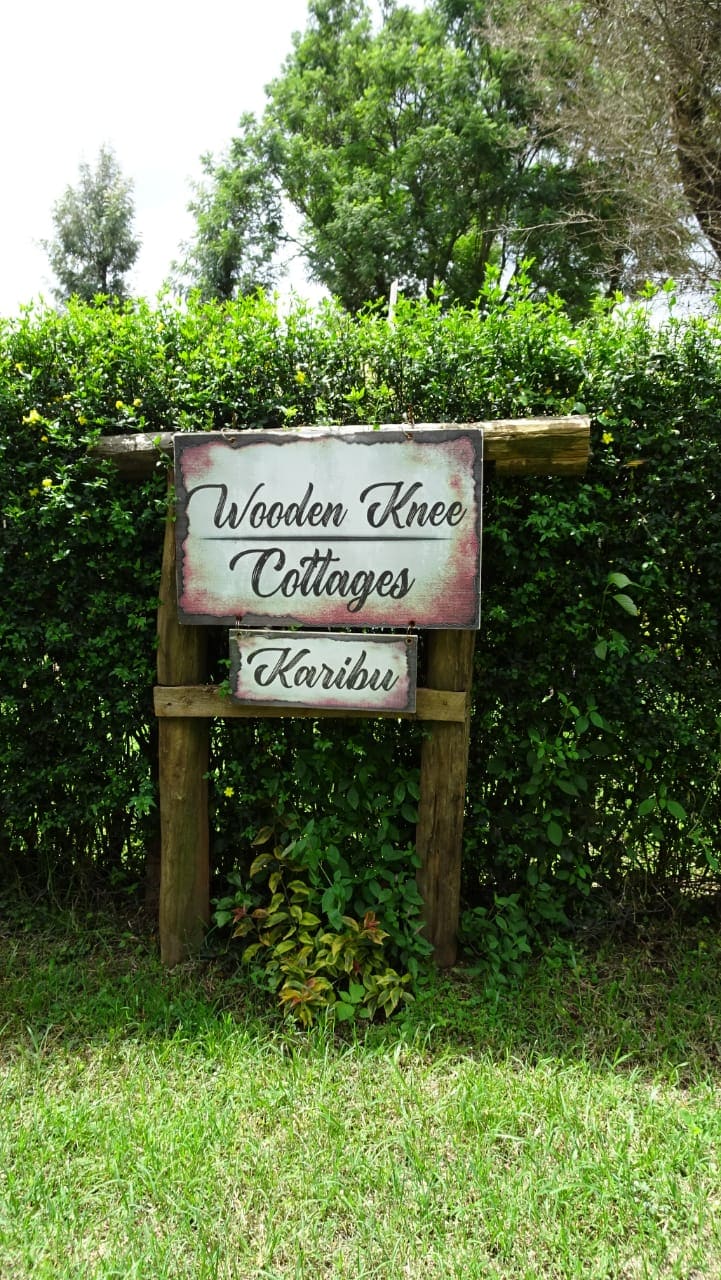 A wooden sign stands at the entrance, displaying 'Wooden Knee Cottages' and 'Karibu' in painted lettering. The sign is surrounded by lush greenery and well-maintained hedges, contributing to a welcoming environment.