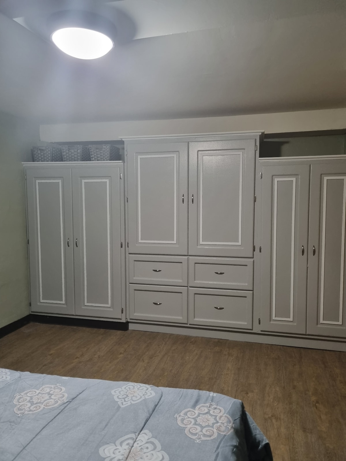A spacious wardrobe occupies one wall, featuring multiple doors and built-in drawers for storage. The light grey finish complements the soft green walls, creating a calm environment. Above the wardrobe, wicker baskets add an organized touch, enhancing the functionality of the room.