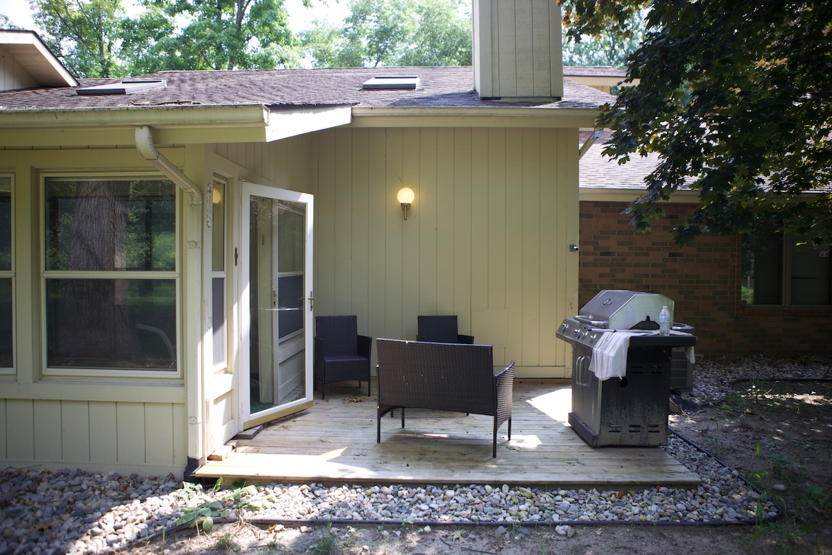 An outdoor patio space features a wooden deck with two dark wicker chairs facing a grill. Natural greenery surrounds the area, creating a quiet retreat for relaxation. A doorway leads into the interior, while soft light illuminates the deck.