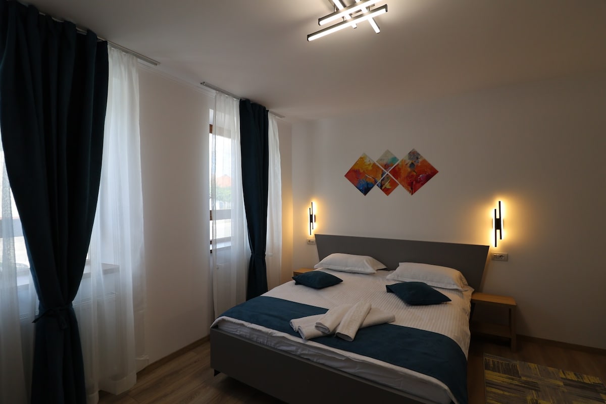 A bright sleeping area features a spacious bed adorned with soft linens and decorative pillows. Two large windows are dressed with sheer curtains, allowing natural light to fill the room. Colorful wall art is displayed above the bed, complemented by modern lighting fixtures.