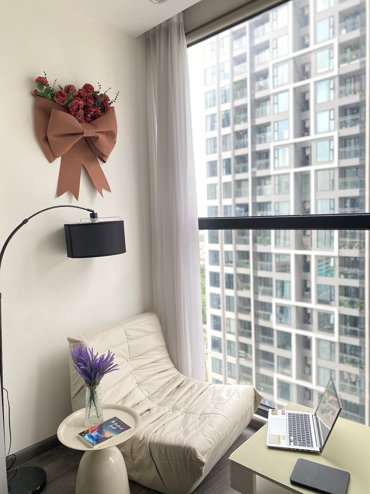 A cozy corner of the room is highlighted by a light-colored armchair facing a window. A small table holds a purple floral arrangement and a book. Stylish decorations include a wall-mounted floral bow, complementing the contemporary backdrop of tall buildings outside.