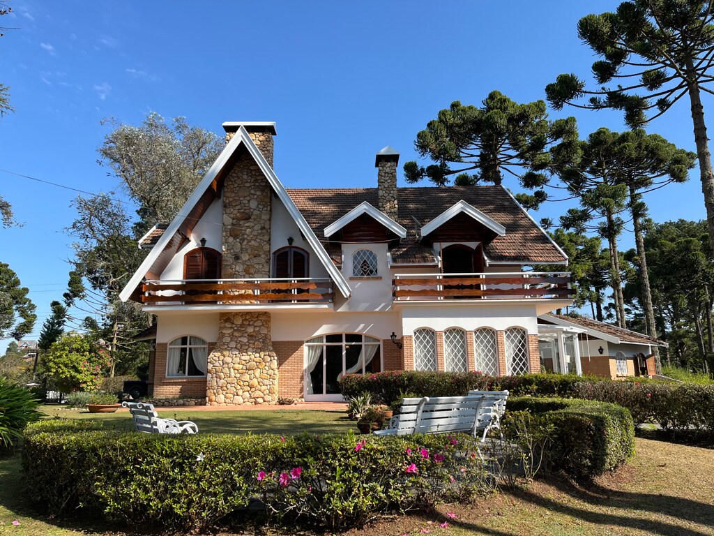 A charming house features a distinct stone and wood exterior, complemented by multiple balconies. Surrounding greenery includes manicured shrubs and garden beds with flowering plants. Clear blue skies enhance the inviting atmosphere, while tall trees stand in the background, providing a sense of natural serenity.