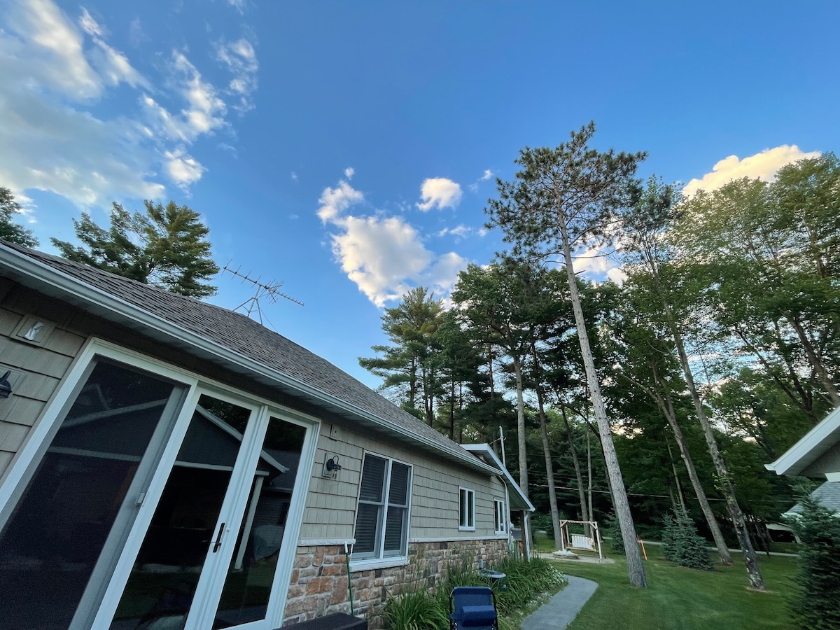 A view of the exterior of a home is presented, showcasing a stone and wood façade under a clear blue sky. Tall trees are visible in the background, providing a natural setting. The structure features multiple windows and a well-maintained pathway leading to the greenery.