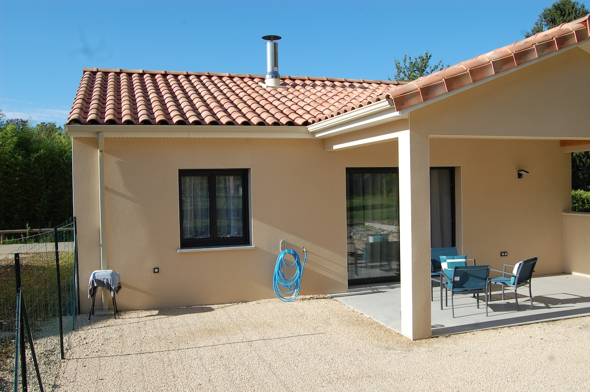 An exterior view of a single-story house is displayed, featuring a terracotta-tiled roof. A small patio area includes modern outdoor seating and a water hose is neatly coiled nearby. Large windows allow natural light to fill the interior, and surrounding greenery is visible.