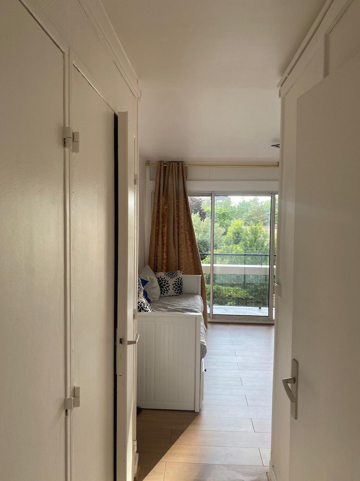 A bright hallway leads to a living area with large glass doors, offering views of greenery outside. Soft beige walls contrast with dark wooden flooring. A cozy seating area, adorned with decorative cushions, invites relaxation and provides access to the balcony.
