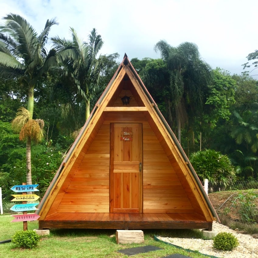 The mini glamping chalé features a distinctive triangular design with wooden cladding, complemented by a warm wooden door. Surrounding greenery includes palm trees and lush vegetation, creating a serene backdrop. Colorful directional signs are positioned nearby, indicating various locations for guests.