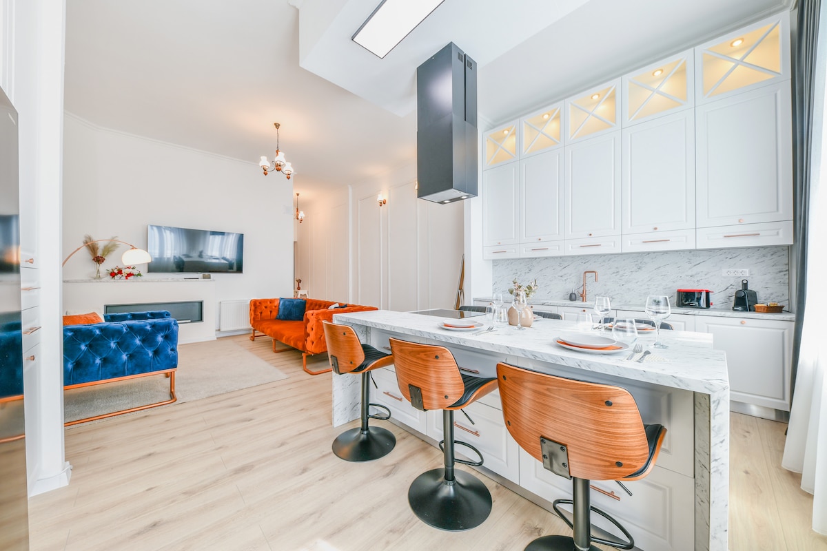 The open kitchen and dining area features modern cabinetry and a marble countertop with bar seating. Stylish stools accompany the kitchen island, while a sleek range hood is positioned above the stovetop. A comfortable seating area with a television is visible in the background.