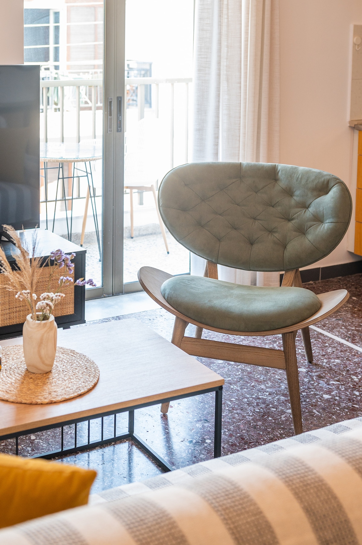 A modern living area features a comfortable green chair with a unique design, positioned adjacent to a wooden coffee table. Natural light enters through a sliding glass door leading to the balcony, enhancing the inviting ambiance of the space.