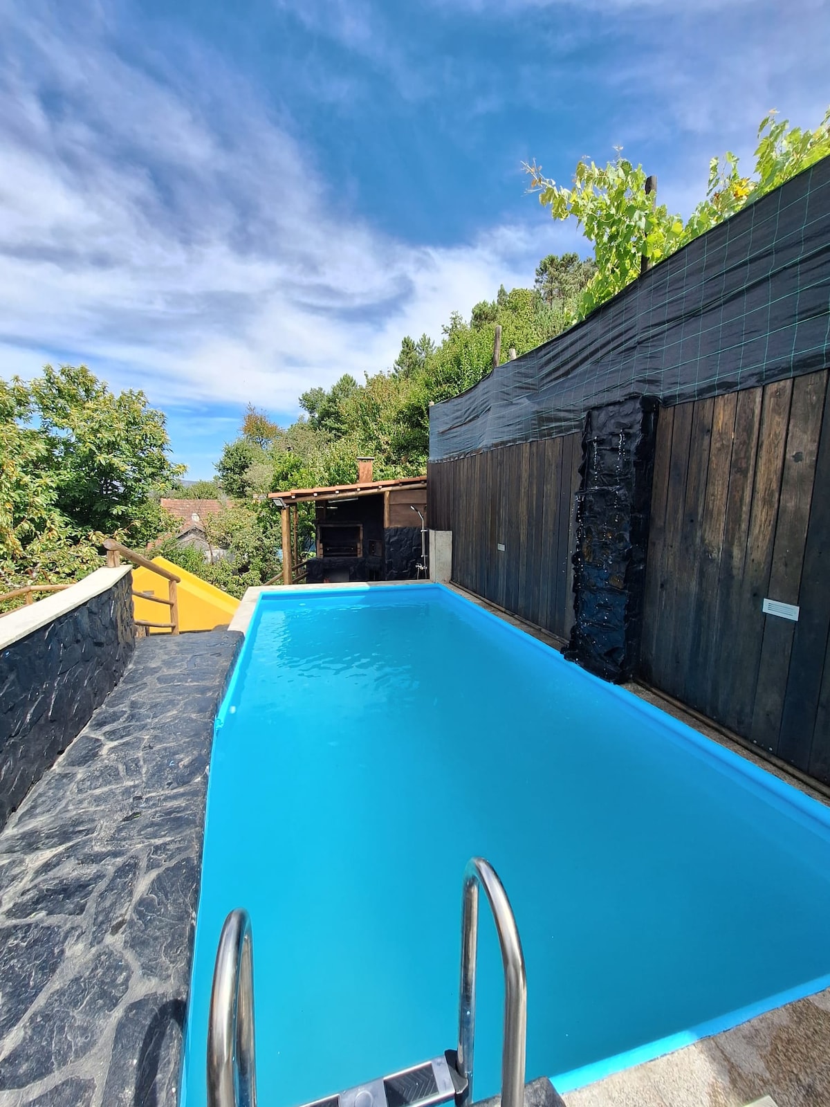 A private swimming pool is surrounded by natural greenery and stone walls. Clear blue water reflects the sky above, while steps lead into the pool. A shaded area is visible in the background, contributing to a serene outdoor atmosphere.