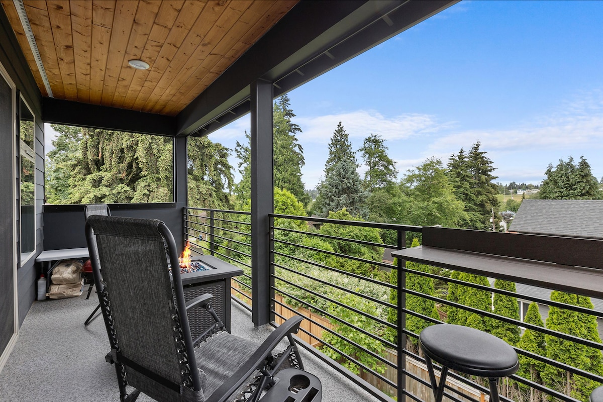 A private balcony offers a fire pit surrounded by comfortable seating. The space features railing views of lush greenery and trees, enhancing the sense of tranquility. The flooring is carpeted, and a small side table is present for convenience.